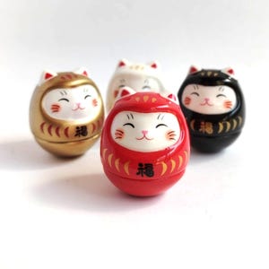 以下が含まれることがあります： 金、白、赤、黒の4体の日本のダルマ猫の置物。それぞれ笑顔の顔、猫の耳、そして「幸運」を表す日本語の文字があります。赤いものは手前にあります。