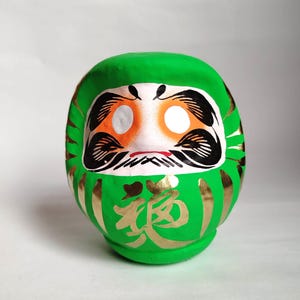 May include: A green Daruma doll with a white face, orange eye sockets, and black details. Gold Japanese characters are painted on the front. The doll is a traditional Japanese good luck charm.