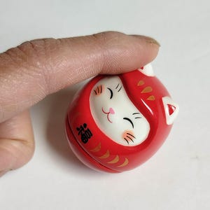 以下が含まれることがあります： 赤いダルマ人形。白い猫の顔、金色のアクセント、そして「幸運」を表す漢字が特徴です。人形は丸く滑らかで、笑顔を浮かべています。