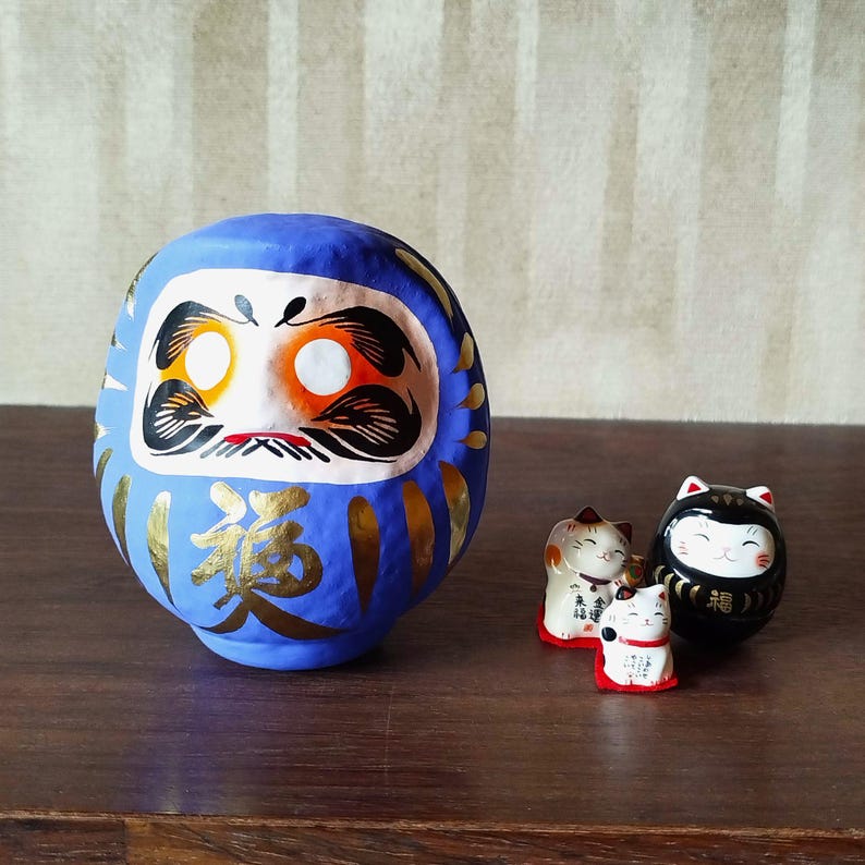 May include: A blue Daruma doll with gold Japanese characters and a detailed face, alongside three small Maneki Neko figurines. The figurines are white, black, and red, with smiling faces and traditional designs. The objects are on a wooden surface.