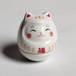 以下が含まれることがあります： 笑顔の猫の顔をした白い陶器のダルマ人形。猫は耳と頬に赤いアクセントがあり、金色のマーキングと赤い「幸運」の漢字があります。