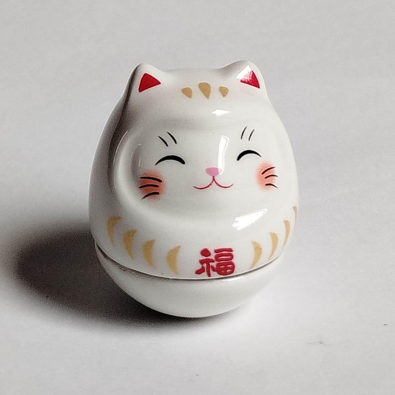 以下が含まれることがあります： 笑顔の猫の顔をした白い陶器のダルマ人形。猫は耳と頬に赤いアクセントがあり、金色のマーキングと赤い「幸運」の漢字があります。