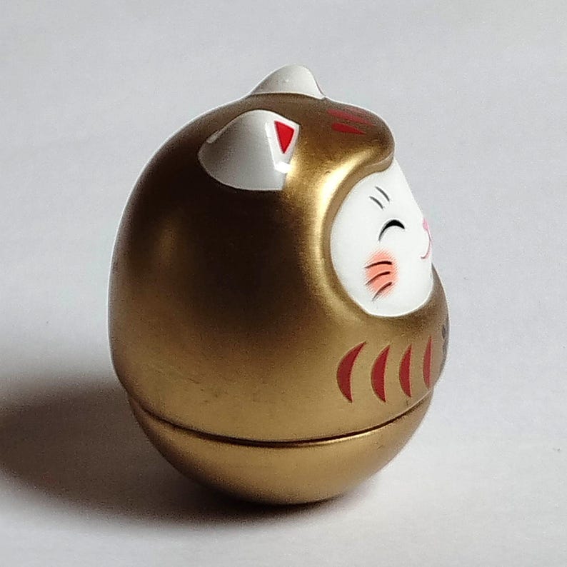 以下が含まれることがあります： 金色のダルマ人形。白い顔、赤いアクセント、笑顔の表情が特徴です。人形は丸い形で、日本の伝統的な幸運のお守りです。人形のデザインには、白い耳と上部の赤いストライプが含まれています。