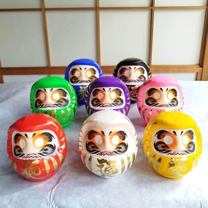 May include: A collection of seven Daruma dolls in various colors, including red, green, blue, purple, black, pink, white, and yellow. Each doll features a detailed face with black facial hair and gold accents. The dolls are arranged on a white surface.
