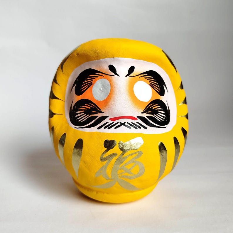 May include: A yellow Daruma doll with black facial features, including eyes, eyebrows, and a mustache. The doll has gold accents and Japanese characters on the front. The Daruma doll is a traditional Japanese good luck charm.