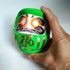 May include: A green Daruma doll with gold accents and Japanese characters. The doll has a white face with orange eye sockets, black eyebrows, and a black mustache. The doll is held in a hand.