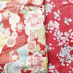 Può includere: Primo piano di due pannelli di tessuto rosso con motivi floreali. Un pannello presenta grandi fiori rosa e bianchi con foglie verdi, mentre l'altro ha delicati fiori di ciliegio bianchi. I tessuti hanno un'estetica tradizionale giapponese.