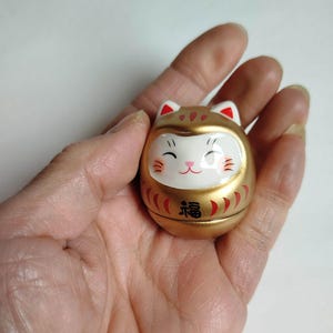 以下が含まれることがあります： 金色のダルマ人形。猫のような顔、赤いアクセント、そして「幸運」を表す日本語の文字が特徴です。人形は白い耳と白い顔をしており、笑顔の表情をしています。人形は手で持たれています。