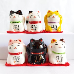 Puede incluir: Seis figuras de cerámica Maneki Neko en varios colores, incluyendo blanco, negro, amarillo y rosa. Cada gato tiene un collar rojo o verde y está sobre una alfombra roja. Texto japonés visible en las figuras.