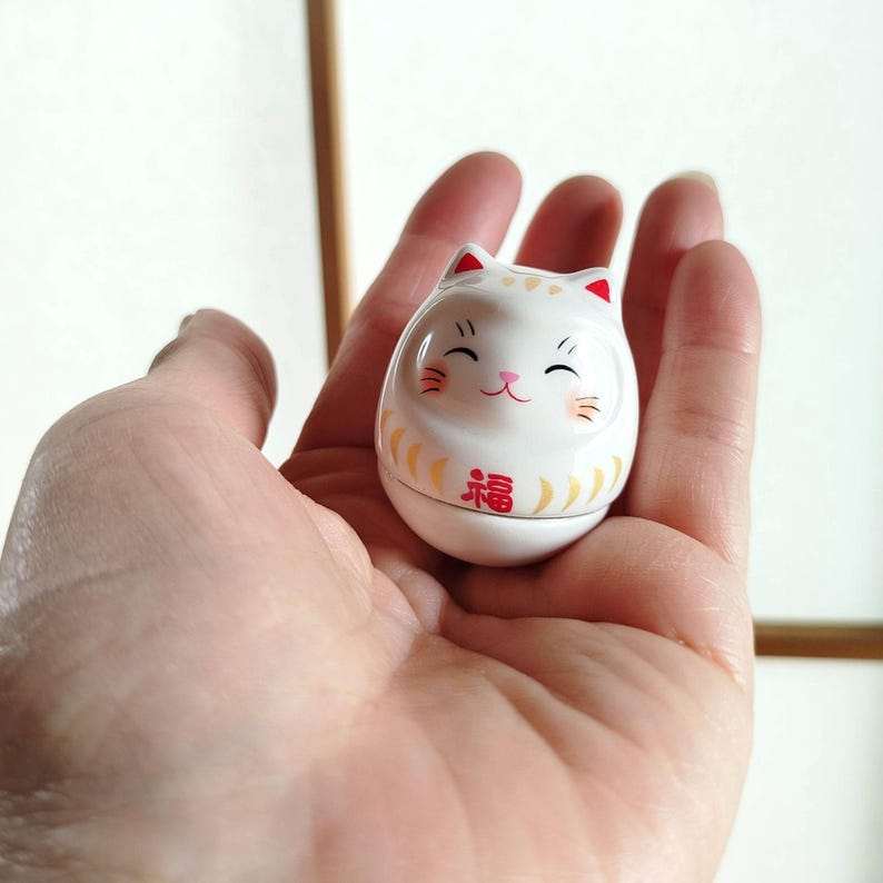 以下が含まれることがあります： 赤い耳と「福」の文字を含む、白いセラミック製の招き猫の置物。 笑顔の猫の置物には金の装飾が施されており、手で持たれています。