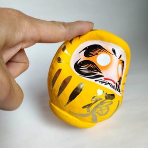 May include: A yellow Daruma doll with gold accents and Japanese calligraphy. The doll has a white face with black and orange details. The Daruma doll is a traditional Japanese good luck charm, often used to set goals.