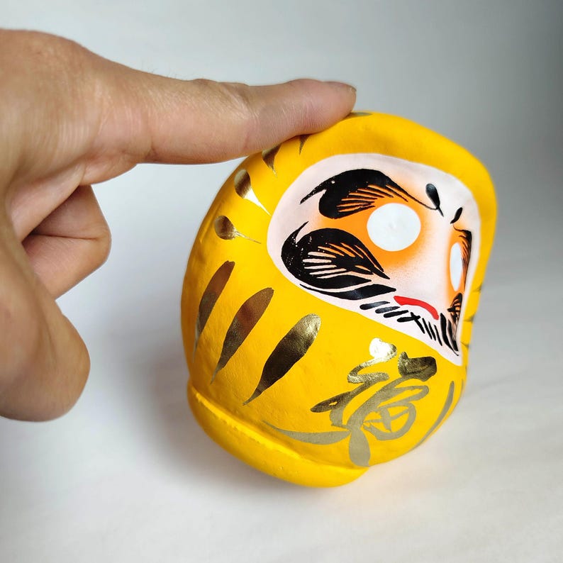 May include: A yellow Daruma doll with gold accents and Japanese calligraphy. The doll has a white face with black and orange details. The Daruma doll is a traditional Japanese good luck charm, often used to set goals.