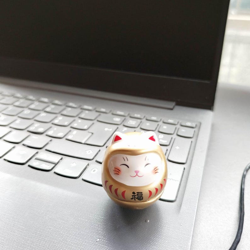 以下が含まれることがあります： 笑顔の猫の顔と赤いアクセントが付いた、小さな金色のダルマ人形。人形の前面には「幸運」を表す日本語の文字があります。人形はラップトップのキーボードの上にあります。