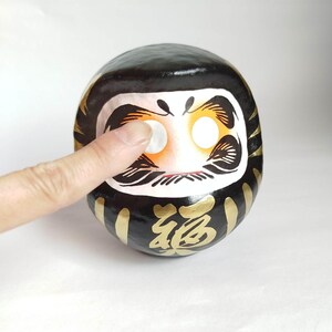 May include: A black Daruma doll with gold accents and the Japanese character for "fortune." The doll has a white face with large, round eyes and a finger touching one eye. The Daruma doll is a traditional Japanese good luck charm.