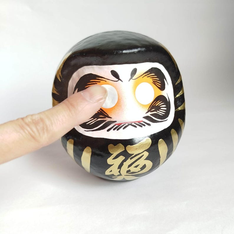 May include: A black Daruma doll with gold accents and the Japanese character for "fortune." The doll has a white face with large, round eyes and a finger touching one eye. The Daruma doll is a traditional Japanese good luck charm.