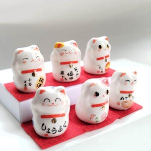 Puede incluir: Seis pequeñas figuras de gato de cerámica blanca, cada una con un collar rojo con un cascabel dorado, expresiones faciales pintadas y caracteres de caligrafía japonesa negra en el frente, dispuestas en dos filas sobre cuadrados de fieltro rojo.