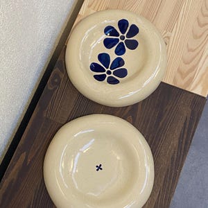 Op de afbeelding: Twee crèmekleurige keramische borden. Eén bord heeft twee blauwe bloemmotieven, de andere een enkel blauw kruis. De borden worden gepresenteerd op een houten oppervlak. Geschikt voor het serveren van voedsel.