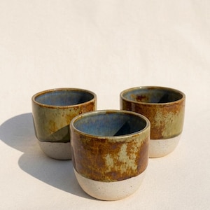 May include: Three handmade ceramic cups with a rustic, textured appearance. The cups have a white base, a brown and green midsection, and a blue interior. The cups are arranged on a white surface, with shadows adding depth.