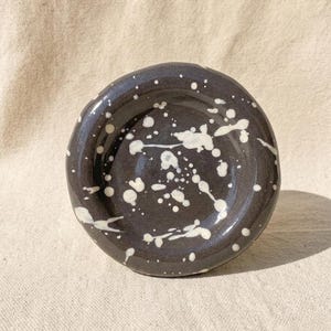 May include: A small, round, handmade ceramic dish with a glossy dark charcoal gray finish. The surface is decorated with an irregular white paint splatter pattern, featuring various sizes of dots and streaks across the entire interior basin.