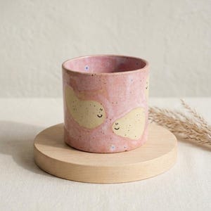 以下が含まれることがあります： 斑点のあるピンク色の釉薬が施された円筒形のセラミックカップ。淡い黄色の豆のような形をした図形が描かれており、黒い線で描かれたシンプルな笑顔が特徴です。カップは滑らかで真っ直ぐな縁と平らな底面を持ち、手作り感のある仕上がりです。