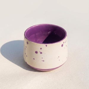 Könnte beinhalten: Kleine, handgefertigte Keramiktasse mit weißer Außenseite und lila Punkten. Innen und am Boden ist sie durchgehend lila. Die runde Form deutet auf die Verwendung für Getränke hin, mit einem Fassungsvermögen von ca. 200 ml.