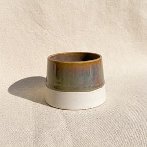 Könnte beinhalten: Eine kleine, handgefertigte Keramik-Tasse mit weißem Boden und glasierter Oberseite. Der obere Bereich wechselt von Grün zu Braun mit schillernden Farbtönen. Die Tasse hat ein schlichtes, modernes Design, ideal für kleine Gegenstände oder als Dekoration.
