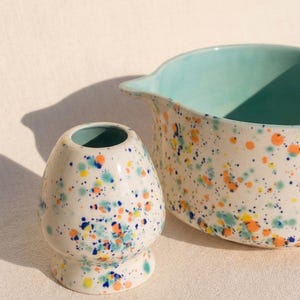 May include: A white ceramic matcha whisk holder and a mixing bowl with a turquoise interior. Both pieces are decorated with colourful splatters of orange, blue, yellow, and green. The whisk holder is egg-shaped and the bowl has a spout.