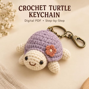 Puede incluir: Un llavero de tortuga de ganchillo con caparazón lavanda, cabeza y extremidades color crema y una pequeña flor naranja. La tortuga tiene ojos de cuentas negras y una boca bordada. Está unida a una anilla y un mosquetón de metal color bronce.