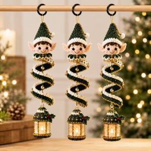 May include: Three hanging crochet elf ornaments, each featuring a green and white pointed hat, a smiling face with pointed ears, and a spiraling green and white body. Each elf holds a small, illuminated lantern decorated with green leaves and red berries.