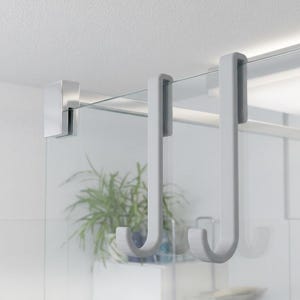 May include: Two matte light grey J-shaped hooks designed to hang over the top edge of a glass shower door. The hooks feature a smooth, uniform finish and a simple, functional design that allows them to slide securely onto the glass panel.