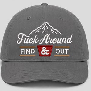 Puede incluir: Una gorra de béisbol gris carbón con texto bordado en blanco que dice "Fuck Around" sobre un gráfico de montaña. Debajo, el texto "FIND & OUT" está bordado en blanco con un borde rectangular rojo alrededor del símbolo ampersand.