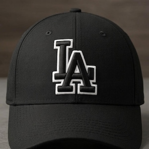 Puede incluir: Gorra de béisbol negra con las letras "LA" bordadas en blanco y negro en la parte delantera. La gorra tiene una visera curva y un botón en la parte superior. El sombrero está hecho de tela duradera.