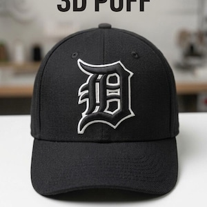 Könnte beinhalten: Schwarze Baseballkappe mit einem weiß-schwarz gestickten "D"-Logo. Die Kappe hat einen gebogenen Schirm und eine strukturierte Krone. Der Text "3D PUFF" steht über der Kappe, und der Text "DST, PES, JEF, EXP, VP3, HUS" steht darunter.