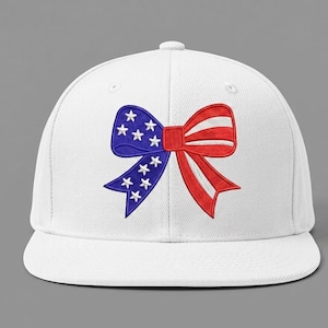 May include: A white baseball cap with an embroidered bow design. The bow is split into two sections: one blue with white stars and the other red with white stripes, creating a patriotic theme.