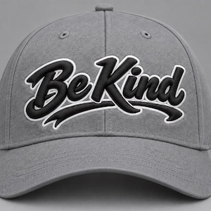 May include: A grey baseball cap featuring the text Be Kind embroidered in a bold, raised black font with a white outline. The cap has a structured crown, a curved brim, and stitched eyelets on the top panels, presenting a clean, uniform appearance.