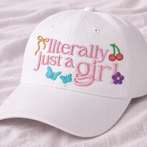 Pode incluir: Boné de beisebol branco com a frase bordada "literally just a girl" em rosa, com um laço amarelo, borboletas azuis, cerejas vermelhas e uma flor roxa. O boné tem uma aba curva e um botão no topo.