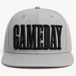 May include: A light gray baseball cap featuring the word GAMEDAY in bold, raised black lettering across the front panels. The cap has a structured crown, a flat brim with stitched detailing, and small ventilation eyelets on the top panels.