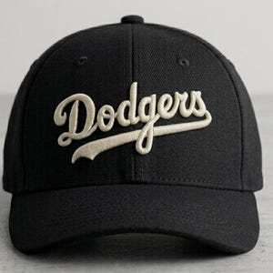 May include: A black baseball cap featuring the word Dodgers embroidered in a cream-colored script font across the front. The cap has a structured crown, a curved brim, and a button on the top center. The embroidery has a raised, textured appearance.