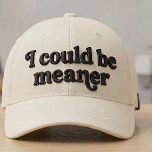 Può includere: Un cappellino da baseball color crema con la scritta "I could be meaner" ricamata in nero a lettere in grassetto sul davanti. Il cappellino ha una visiera curva e un bottone sulla parte superiore. Altri cappellini sono visibili sullo sfondo.