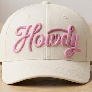 May include: A cream-colored baseball cap featuring the word Howdy embroidered in raised, puffy pink lettering across the front panels. The cap has a structured crown, a curved brim, and matching cream-colored stitching throughout the fabric.