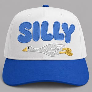 May include: White and blue baseball cap with the word "SILLY" embroidered in blue bubble letters. A cartoon goose is embroidered below the word. The cap has a blue brim and a blue button on top.