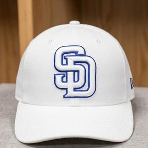 May include: A white baseball cap featuring the San Diego Padres logo in blue. The logo is an interlocking "SD" design. The cap has a curved brim and a structured crown. The New Era logo is visible on the side.