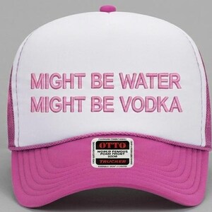 May include: A white and pink trucker hat with the text "MIGHT BE WATER MIGHT BE VODKA" embroidered in pink. The hat has a pink brim and a pink button on top. The hat has a mesh back and a small tag on the front.