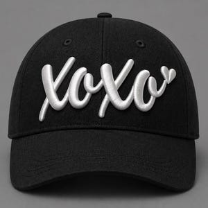 以下が含まれることがあります： 白い筆記体で「XOXO」の刺繍が施された黒い野球帽。帽子にはカーブしたつばと上部にボタンが付いています。テキストはハグとキスの様式化された表現です。