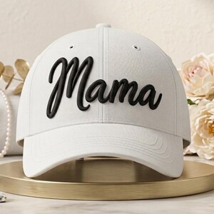 Könnte beinhalten: Eine weiße Baseballkappe mit dem Wort Mama in schwarzer Schreibschrift auf der Vorderseite. Die Kappe hat eine strukturierte Krone, einen gebogenen Schirm und Belüftungsösen an den oberen Paneelen und zeigt ein schlichtes Design.