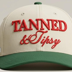May include: A cream-coloured baseball cap with a green peak. The words "TANNED & Tipsy" are embroidered in red on the front. The cap has a curved peak and a button on top.