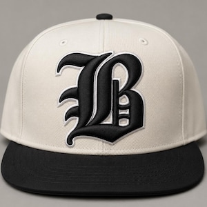 Op de afbeelding: Een baseballpet met een gebroken witte kroon en een zwarte, platte klep. Het voorpaneel is voorzien van een grote, geborduurde letter B in een zwart gotisch lettertype met een witte omlijning. De knoop aan de bovenkant is zwart.