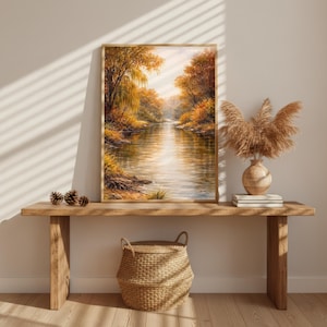 Könnte beinhalten: Ein gerahmtes Landschaftsgemälde zeigt eine ruhige Flussszene mit Herbstlaub in Gold-, Orange- und Brauntönen. Das Kunstwerk wird auf einem Holztisch ausgestellt, ergänzt durch einen geflochtenen Korb, eine dekorative Vase und getrocknete Pflanzen.
