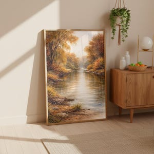 May include: A framed landscape painting depicts a serene river scene. The artwork showcases a river flowing through a forest with trees in autumn colors. The painting is in a wooden frame and leans against a wall.