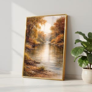 May include: A framed landscape painting depicts a serene river scene. The artwork features a golden frame and showcases a river flowing through a forest with autumn foliage in shades of orange, yellow, and brown. The painting is leaning against a white wall.